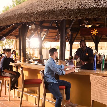 Amwaj Rotana, Jumeirah Beach - Dubai Exterior photo The photo shows a bar area with a thatched roof. There are several people seated at the bar. On the left, two individuals are chatting together, while a bartender is serving a customer in the foreground. The customer is wearing a blue shirt and sitti