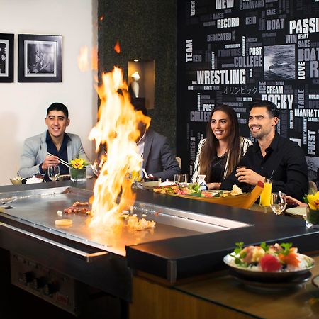Amwaj Rotana, Jumeirah Beach - Dubai Exterior photo The photo shows a lively dining scene at a restaurant where hibachi cooking is taking place. In the foreground, a chef is preparing food on a grill, creating flames, which adds excitement to the experience. There are five diners seated around the gri