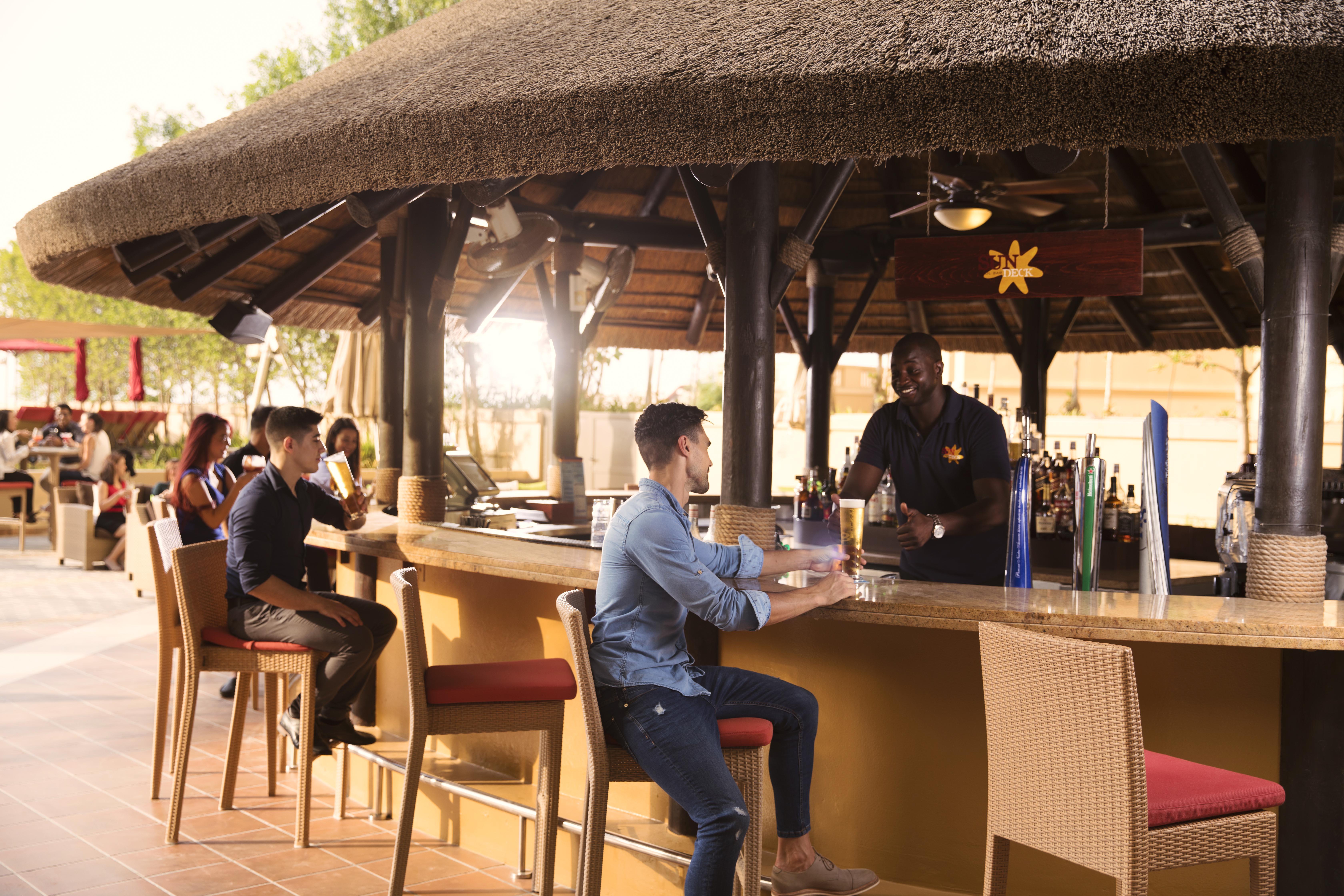 Amwaj Rotana, Jumeirah Beach - Dubai Exterior photo The photo shows a bar area with a thatched roof. There are several people seated at the bar. On the left, two individuals are chatting together, while a bartender is serving a customer in the foreground. The customer is wearing a blue shirt and sitti