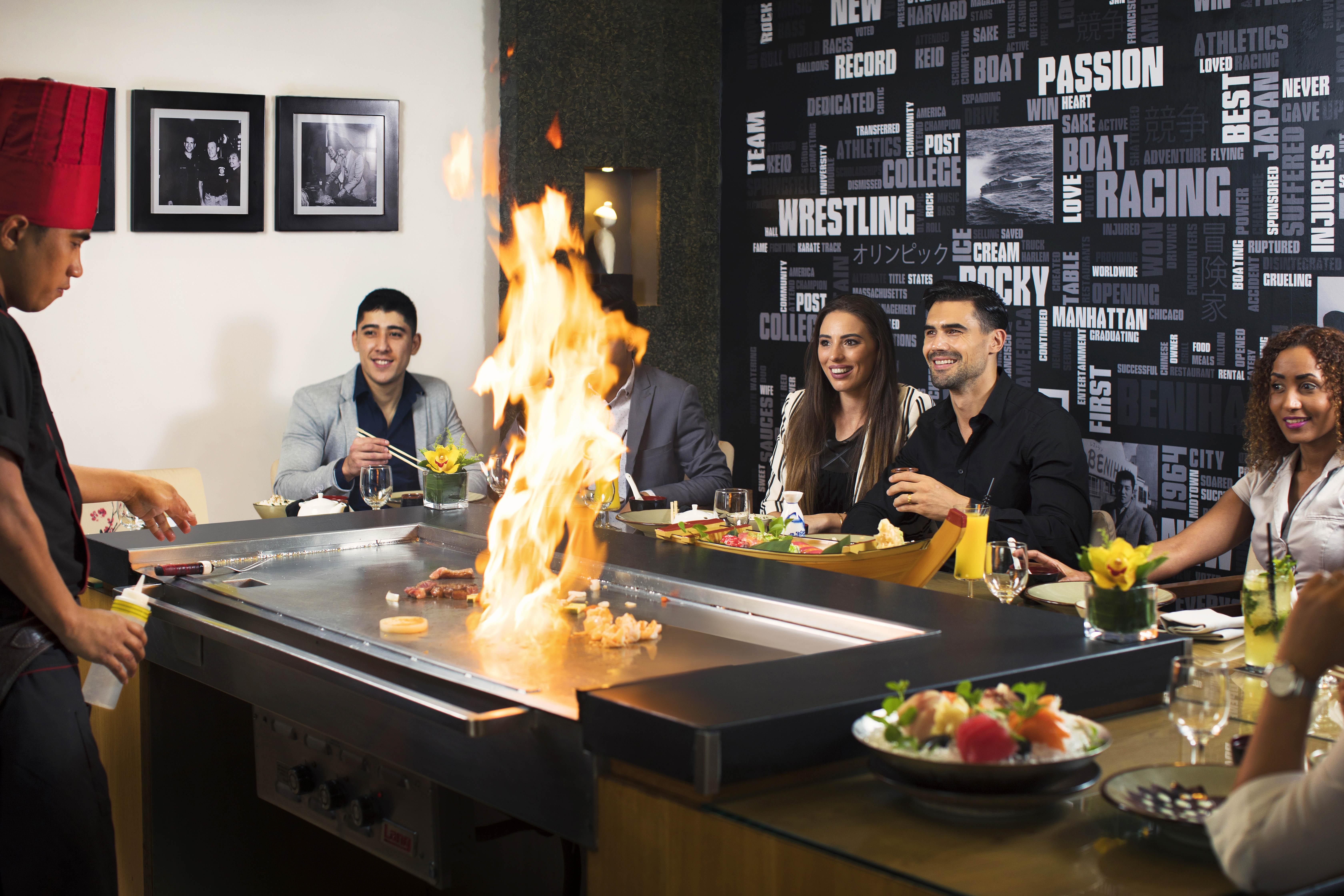 Amwaj Rotana, Jumeirah Beach - Dubai Exterior photo The photo shows a lively dining scene at a restaurant where hibachi cooking is taking place. In the foreground, a chef is preparing food on a grill, creating flames, which adds excitement to the experience. There are five diners seated around the gri