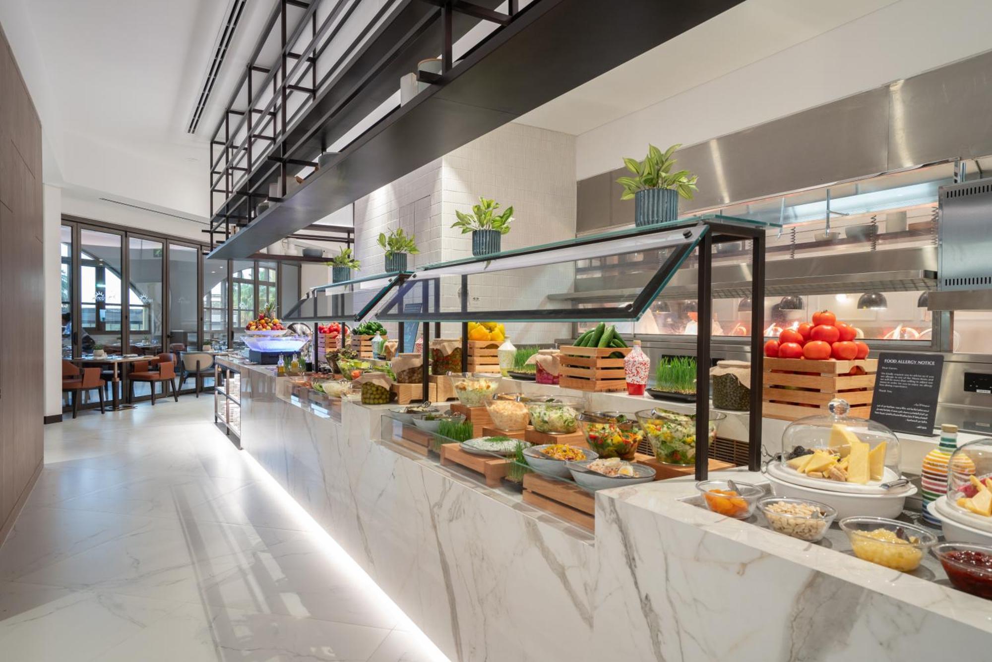 Amwaj Rotana, Jumeirah Beach - Dubai Exterior photo The photo depicts a modern, stylish buffet area in a restaurant. It features a long marble countertop with an array of fresh ingredients and food items displayed attractively. There are different types of vegetables, fruits, cheeses, and other dishes