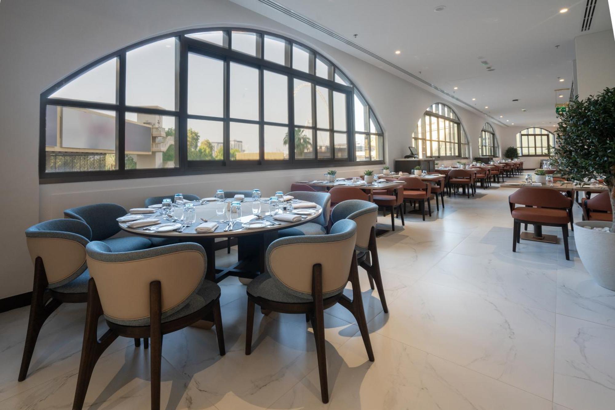 Amwaj Rotana, Jumeirah Beach - Dubai Exterior photo The photo shows the interior of a modern restaurant. It features a spacious layout with several tables arranged around the room. The design includes elegant, upholstered chairs with a mix of colors and round tables set with plates and cutlery. Large 