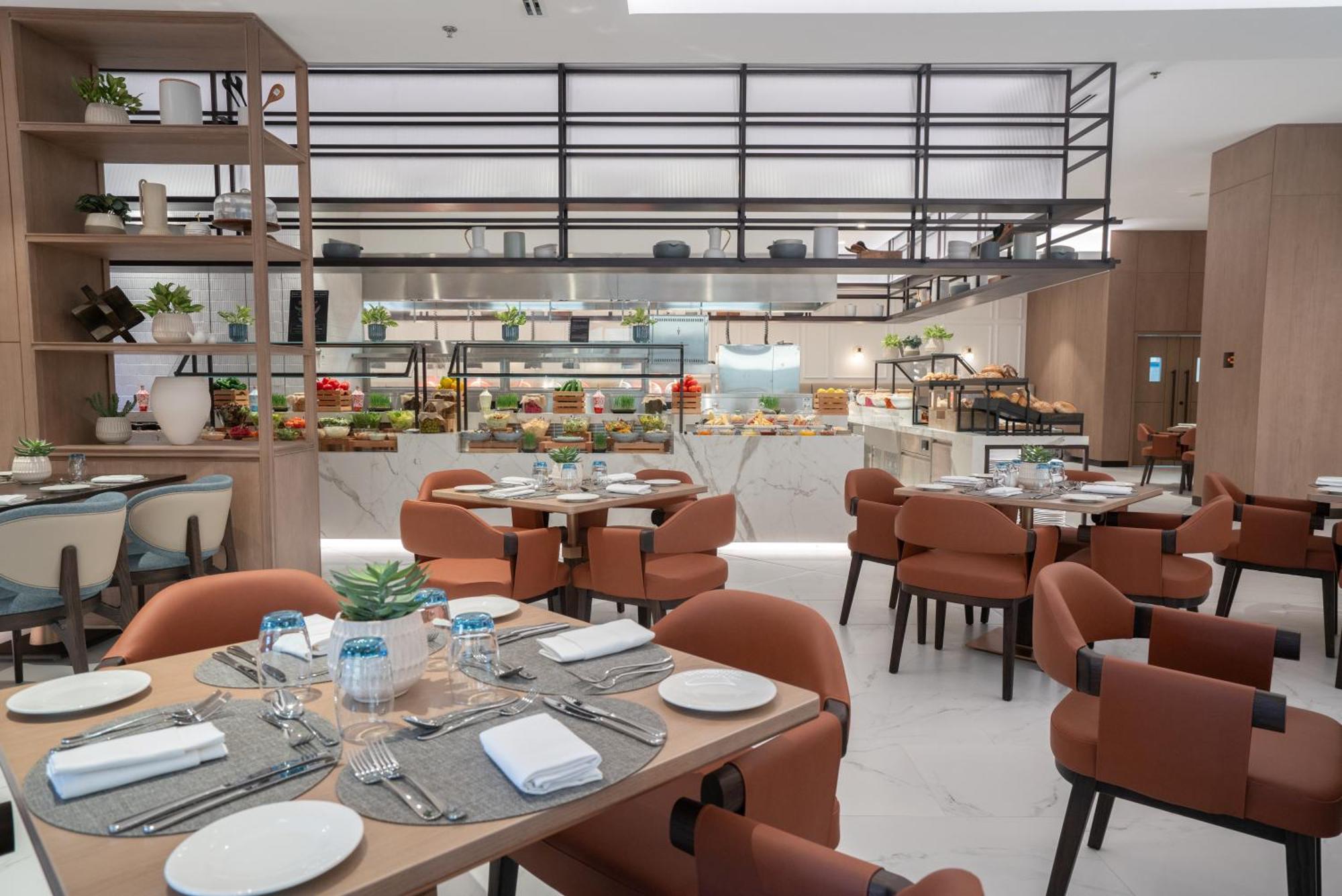 Amwaj Rotana, Jumeirah Beach - Dubai Exterior photo The photo shows a modern restaurant interior with a clean and stylish design. In the foreground, there are several tables set for dining, each with plates, cutlery, and glasses. The chairs are a warm rust color, creating a cozy atmosphere. 

In the b