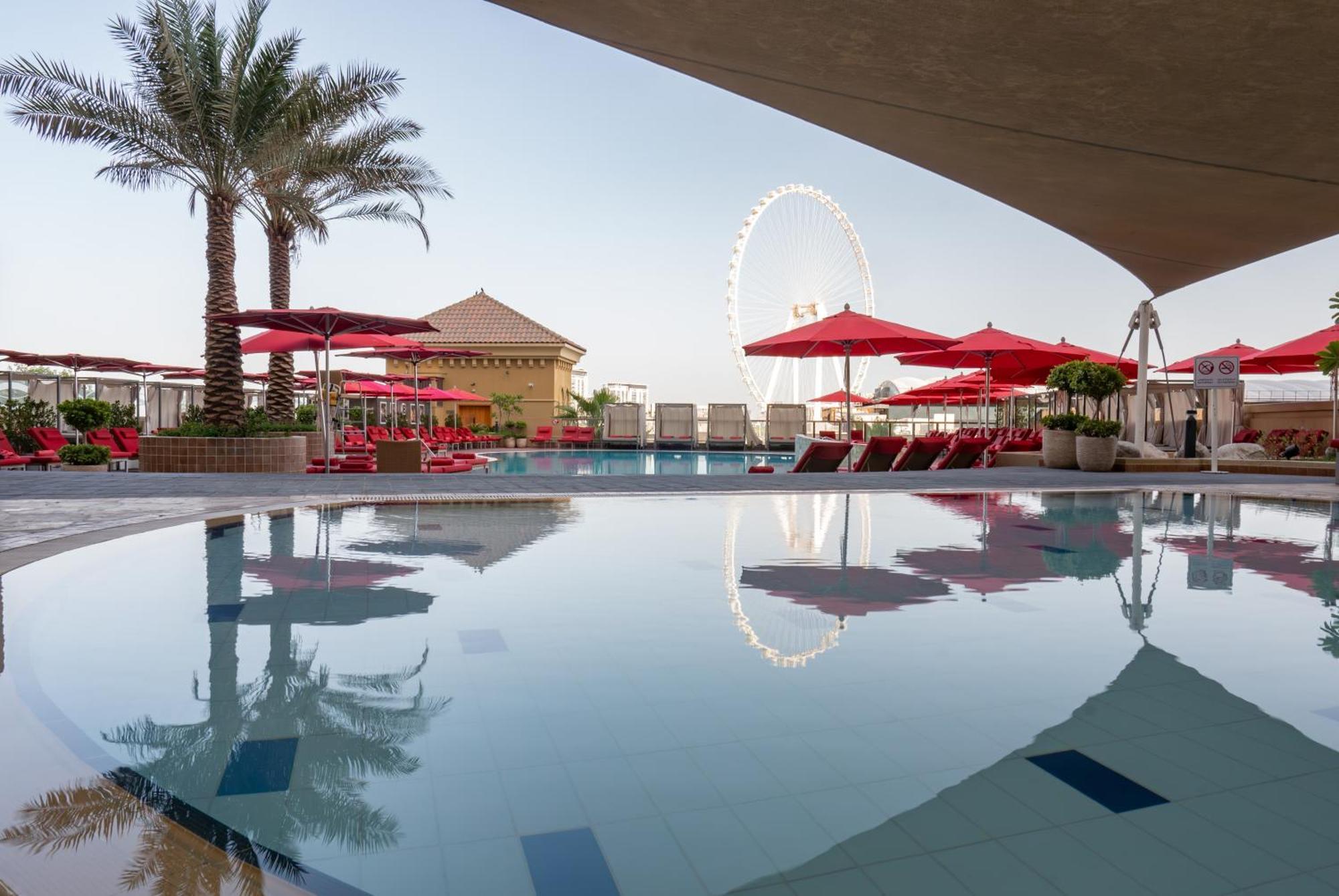 Amwaj Rotana, Jumeirah Beach - Dubai Exterior photo The photo depicts a luxurious outdoor pool area surrounded by palm trees and lounge chairs. There are several red umbrellas set up near the poolside, creating a vibrant and inviting atmosphere. In the background, there's a large wheel—likely a Ferris