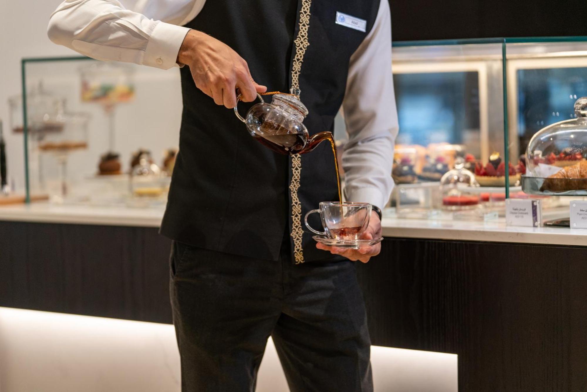 Amwaj Rotana, Jumeirah Beach - Dubai Exterior photo The photo shows a person in a black vest and white shirt pouring a beverage from a glass teapot into a clear glass cup. The setting appears to be a café or restaurant, with a display case in the background containing various pastries or desserts. The