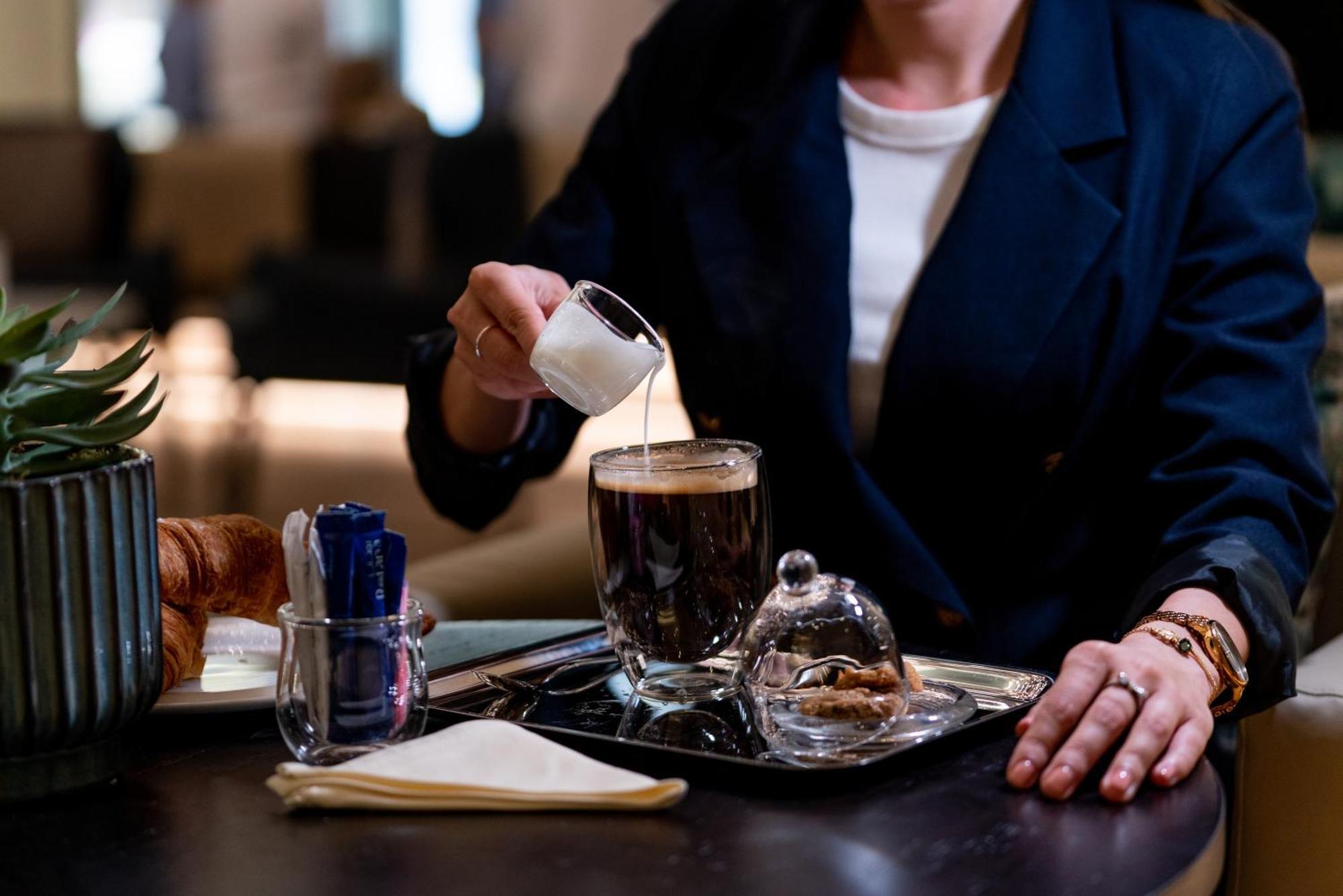 Amwaj Rotana, Jumeirah Beach - Dubai Exterior photo The photo shows a person pouring cream from a small pitcher into a cup of coffee. The coffee is on a tray, which also holds a glass dome covering some pastries or cookies. There are utensils in a holder, a folded napkin, and a croissant visible on th