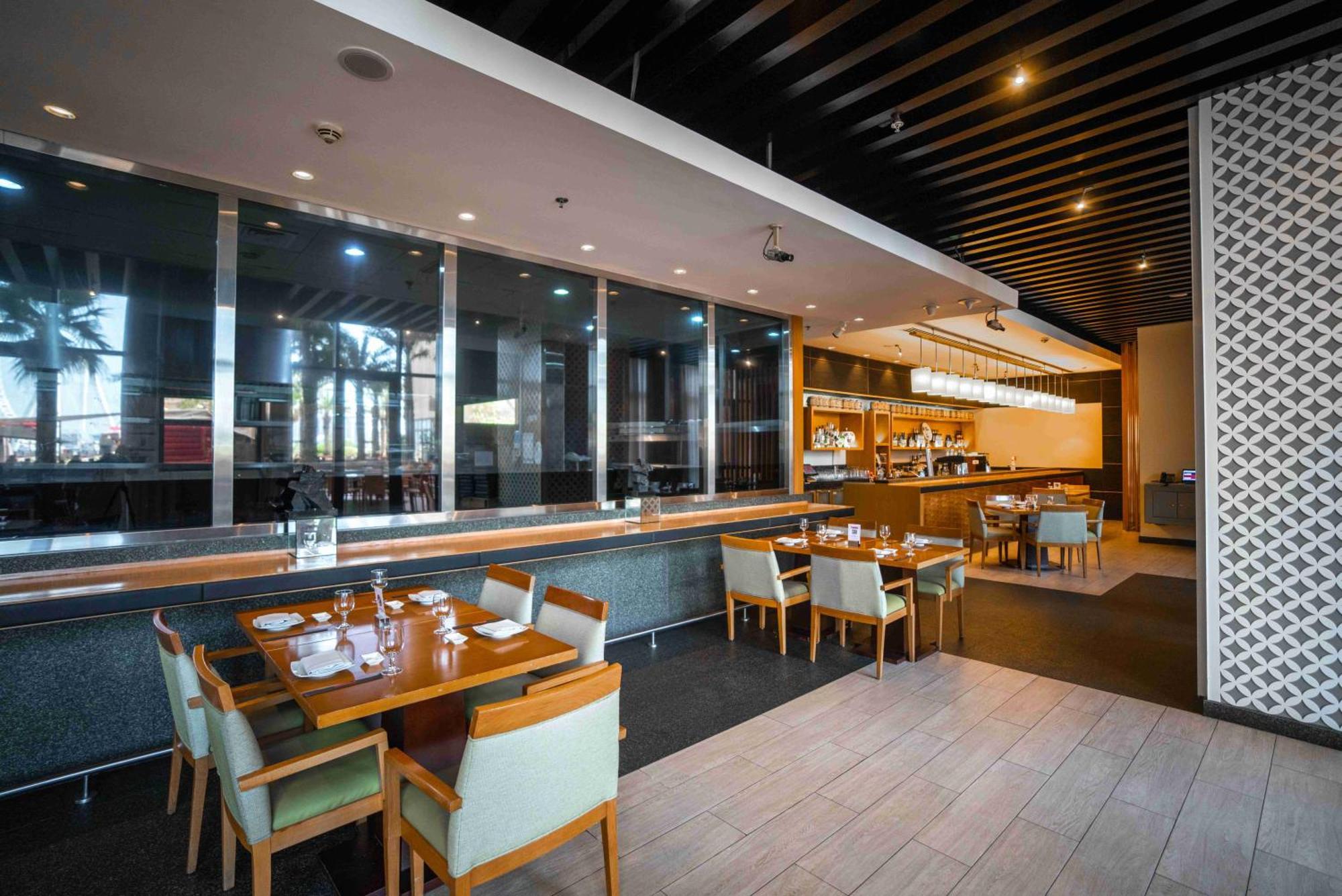 Amwaj Rotana, Jumeirah Beach - Dubai Exterior photo The photo shows the interior of a modern restaurant. In the foreground, there are several wooden tables set with utensils, plates, and glassware. The chairs are upholstered in a light fabric. The background features a counter area with a coffee machi