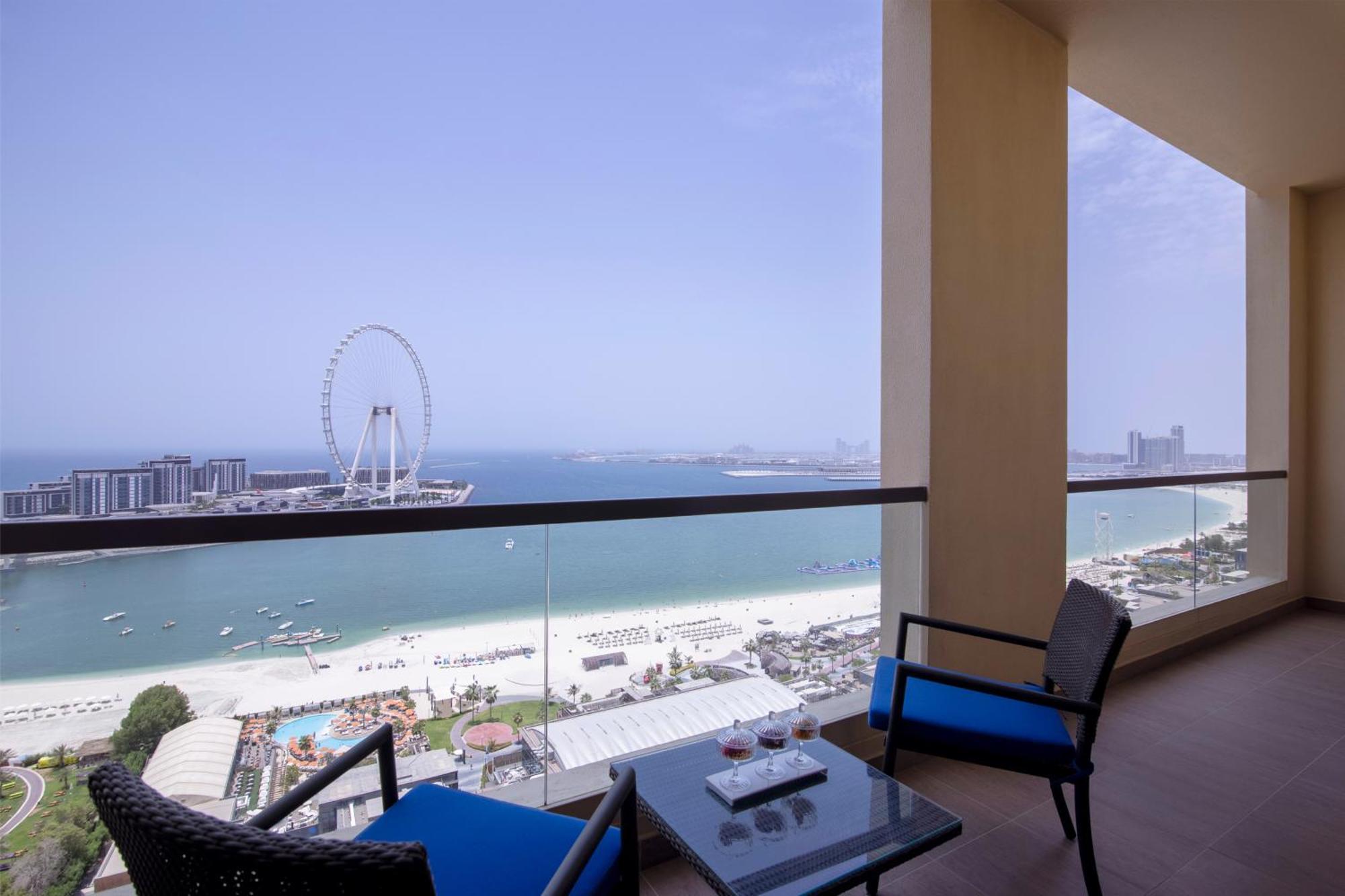 Amwaj Rotana, Jumeirah Beach - Dubai Exterior photo The photo depicts a balcony scene overlooking a beach and sea. There's a modern, stylish balcony with two blue chairs and a small glass table. In the background, you can see a wide expanse of sandy beach, water, and a large Ferris wheel. The sky is c